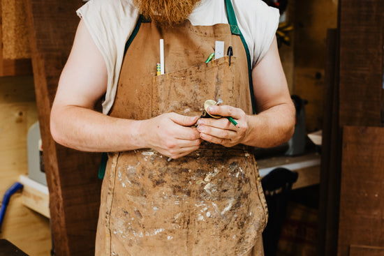 A bearded carpenter holding a scribe and a pencil in front of his brown apron. The apron is covered in old paint stains.