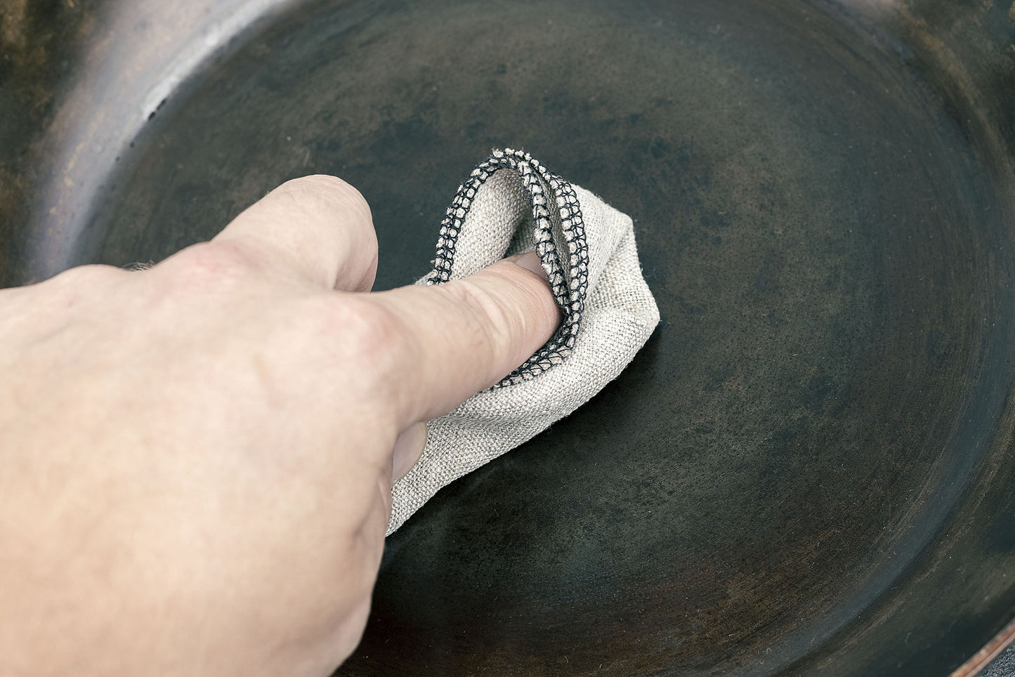 A hand holding a bunched up linen cloth applying a thin layer of seasoning paste to a carbon steel skillet.